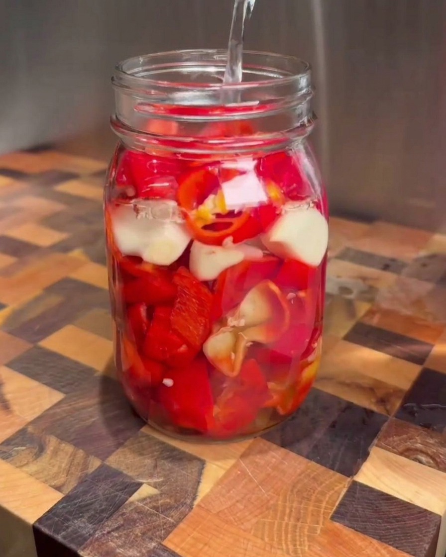Klare Salzlake wird aus einem Messbecher in ein Einmachglas voller gehackter roter Chilis und Knoblauchzehen gegossen.