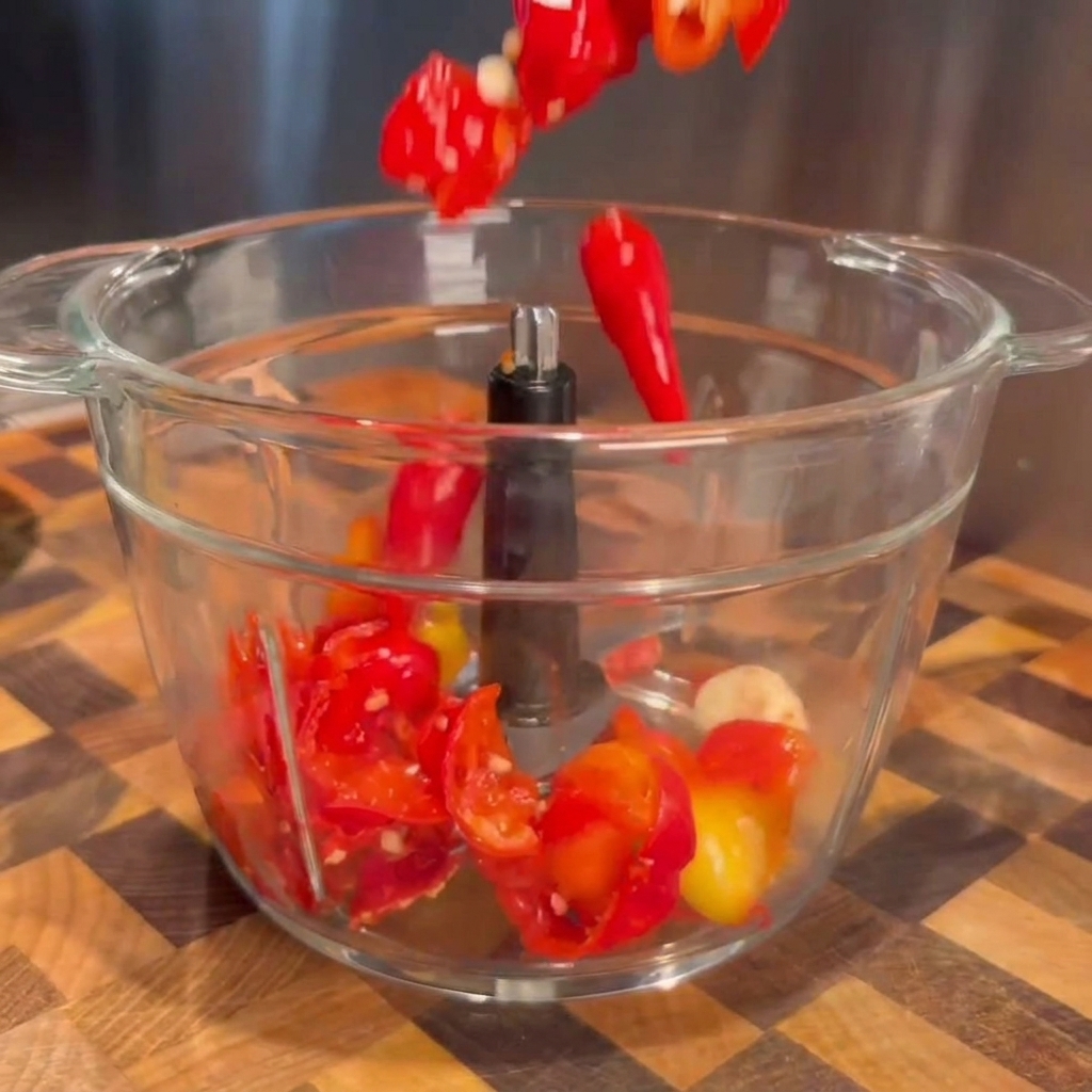 Abgetropfte fermentierte rote Chilis und Knoblauchzehen fallen in den Behälter einer Küchenmaschine.