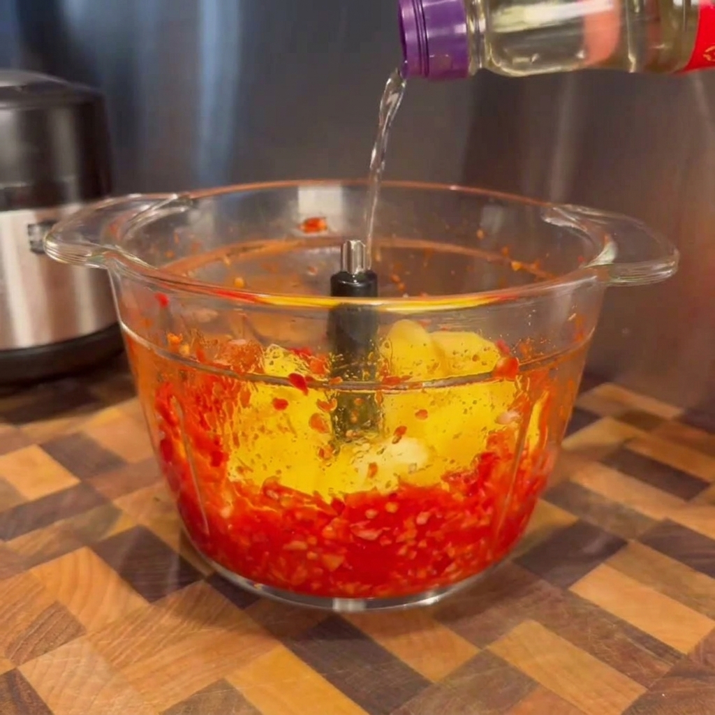Eine klare Flüssigkeit wird aus einer Flasche in den Mixer gegossen, in dem sich bereits die pürierten roten Chilis und Mangostücke befinden.