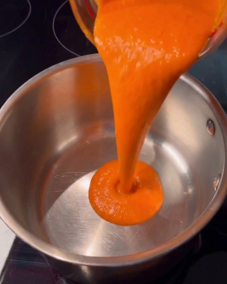 Leuchtend orange Hot Sauce wird gleichmäßig aus einem Glasgefäß in einen sauberen Edelstahltopf auf dem Herd gegossen.