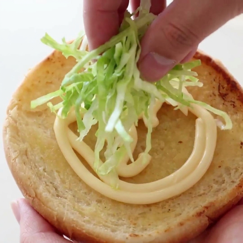 Eine Hand legt einen Haufen knackigen, fein geschnittenen grünen Salat auf ein getoastetes Burgerbrötchen mit Mayonnaise.