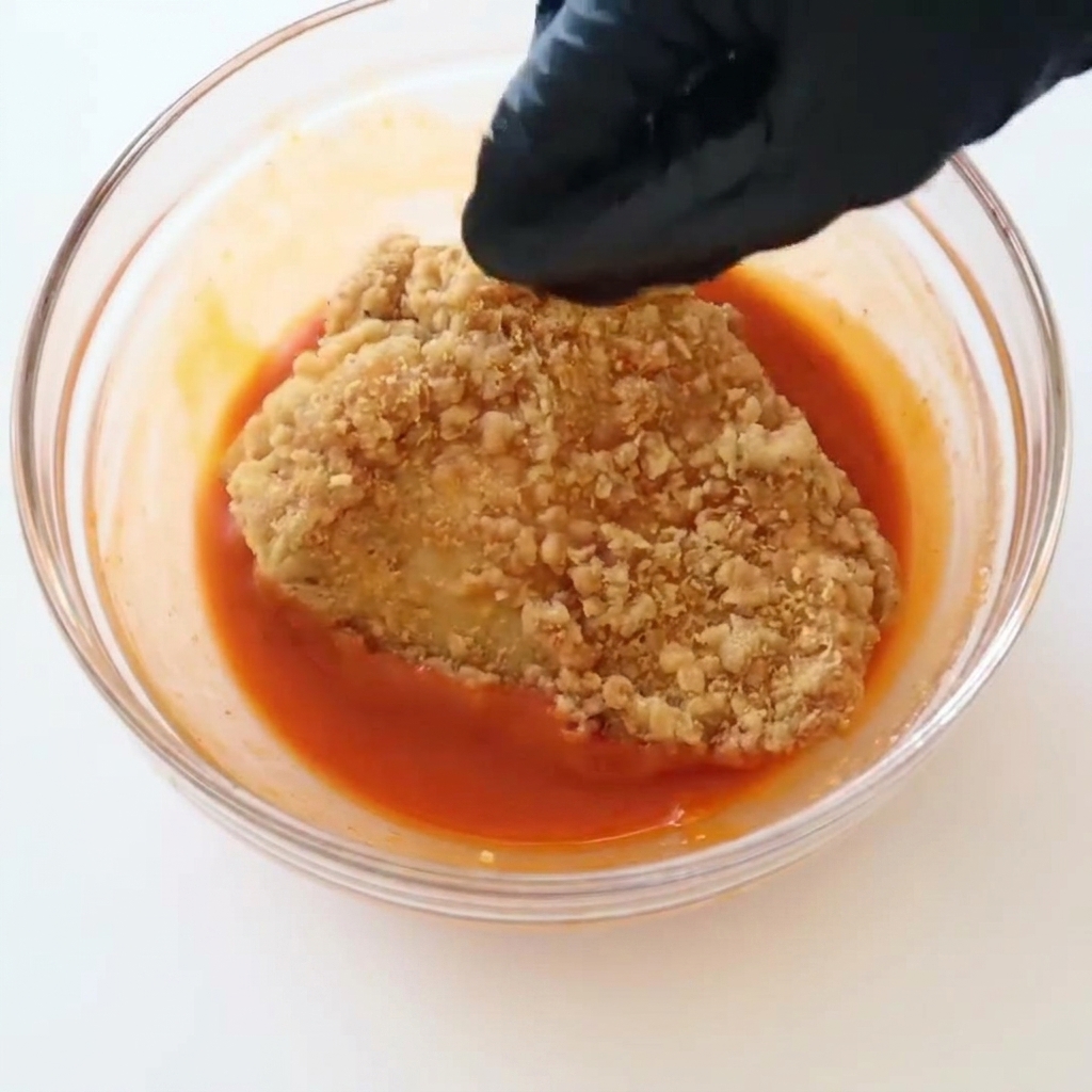 Eine Hand mit schwarzem Handschuh taucht ein knuspriges, goldbraun frittiertes Hähnchenfilet in eine Schüssel mit leuchtend roter Buffalo-Sauce.