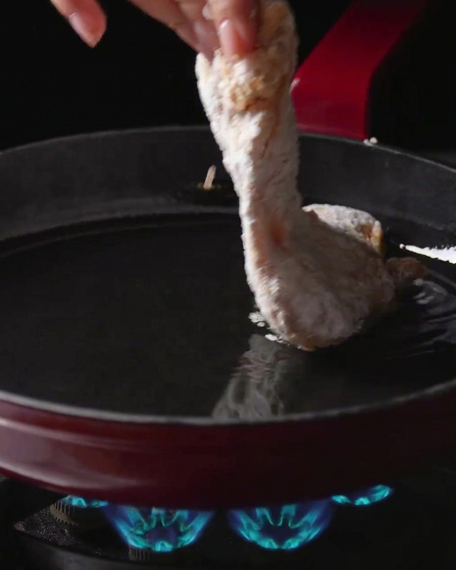 Eine Hand legt eine mit Stärke bestäubte Hähnchenkeule in eine Pfanne mit heißem Öl über einer brennenden Gasflamme.