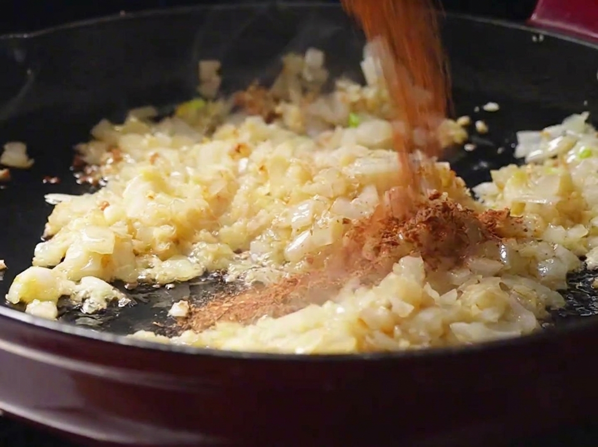 Hausgemachtes rotbraunes Cajun-Gewürzpulver wird über angedünstete Zwiebeln und Knoblauch in einer Bratpfanne gestreut.