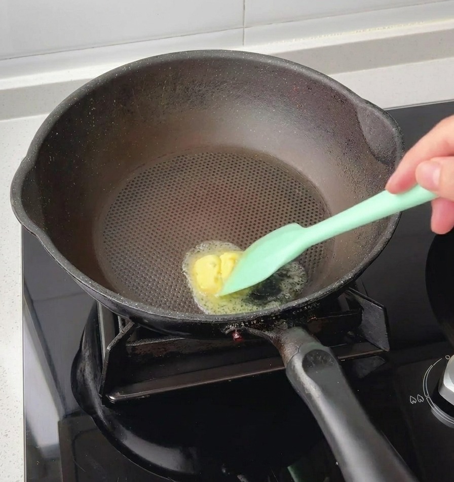 Ein grüner Silikonspatel lässt ein kleines Stück gelbe Butter in der Mitte einer heißen schwarzen Pfanne schmelzen.
