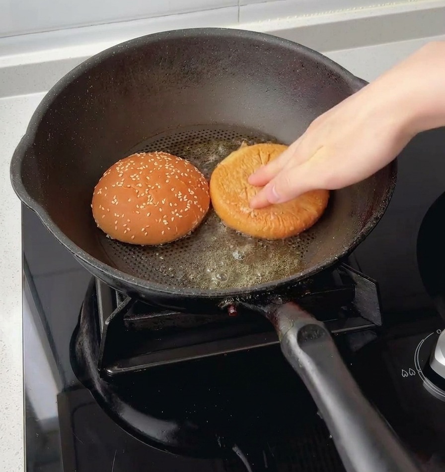 Eine Hand drückt die oberen und unteren Hälften eines Sesam-Burgerbrötchens mit der Schnittfläche nach unten in geschmolzene Butter in einer Pfanne.