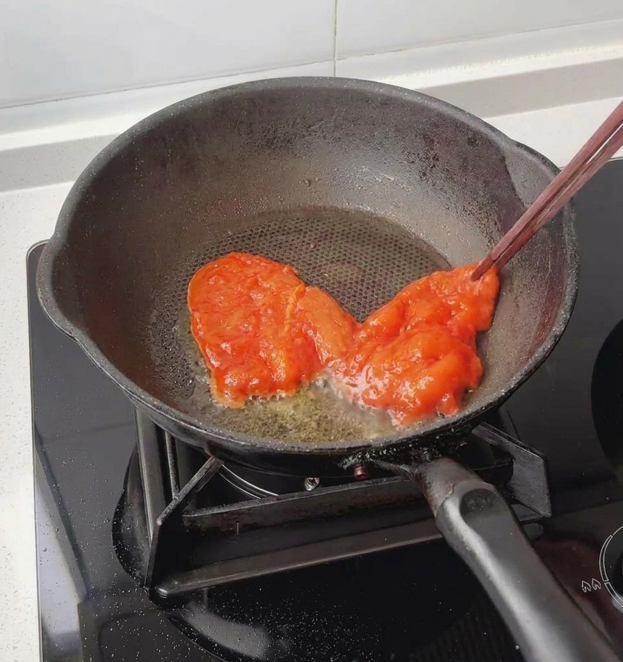 Mit Stäbchen wird eine rote, fertig marinierte Hähnchenbrust in eine leicht gefettete schwarze Bratpfanne gelegt.