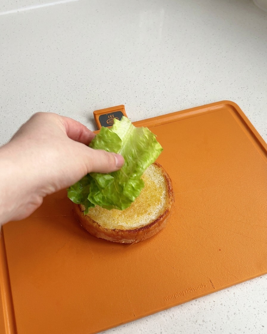 Eine Hand legt ein sauberes, knackig grünes Salatblatt auf die Unterseite eines goldbraun in Butter gerösteten Burgerbrötchens auf einem orangenen Schneidebrett.