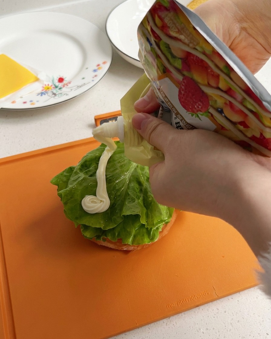 Cremige weiße Mayonnaise wird in einer dicken Linie über ein grünes Salatblatt auf der Brötchenunterseite gegeben.
