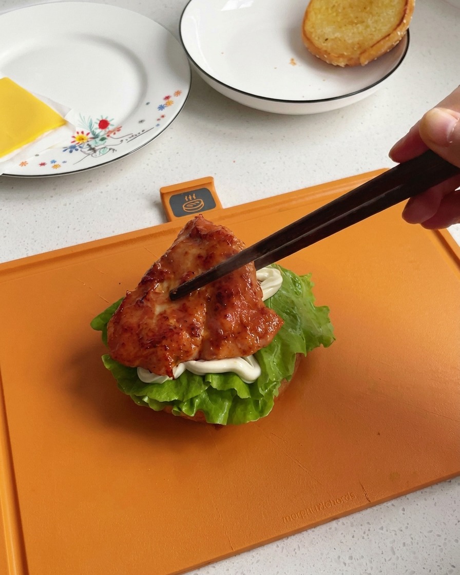 Eine Hand legt mit dunklen Holzstäbchen ein glänzendes, karamellisiertes Stück gebratene Hähnchenbrust auf den Burger-Stapel.