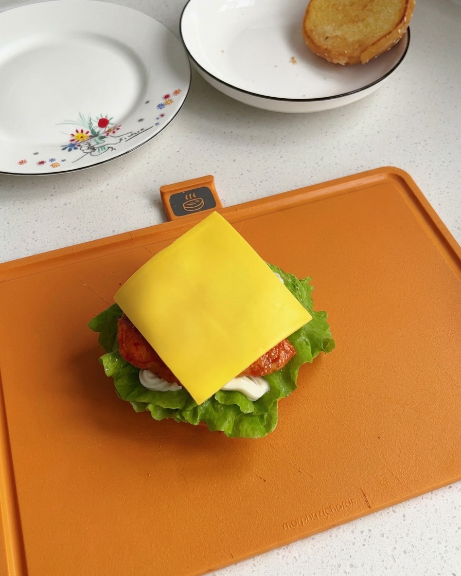 Eine quadratische Scheibe hellgelber Schmelzkäse liegt genau auf einem frisch gebratenen Stück Hähnchen im unvollständigen Burger.