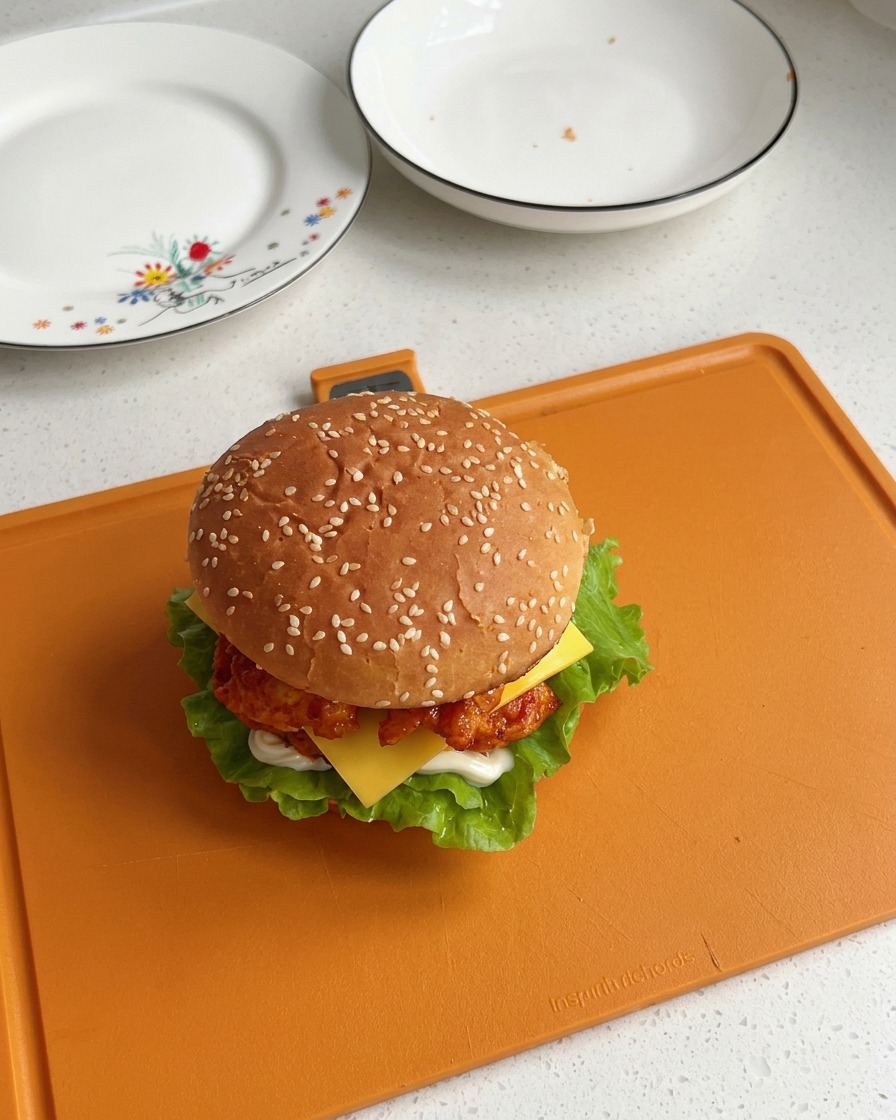 Ein vollständig zusammengesetzter Orleans Chicken Burger mit Sesambrötchen zeigt sichtbare Schichten von Salat, Hähnchen und leicht geschmolzenem Käse.