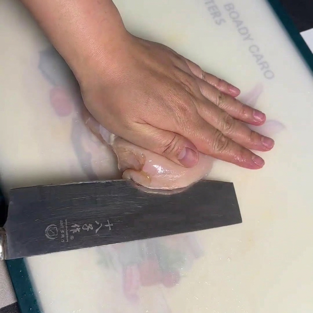 Eine Hand hält eine rohe Hähnchenbrust flach auf einem weißen Schneidebrett, während sie mit einem großen Kochmesser vorsichtig horizontal durchgeschnitten wird.