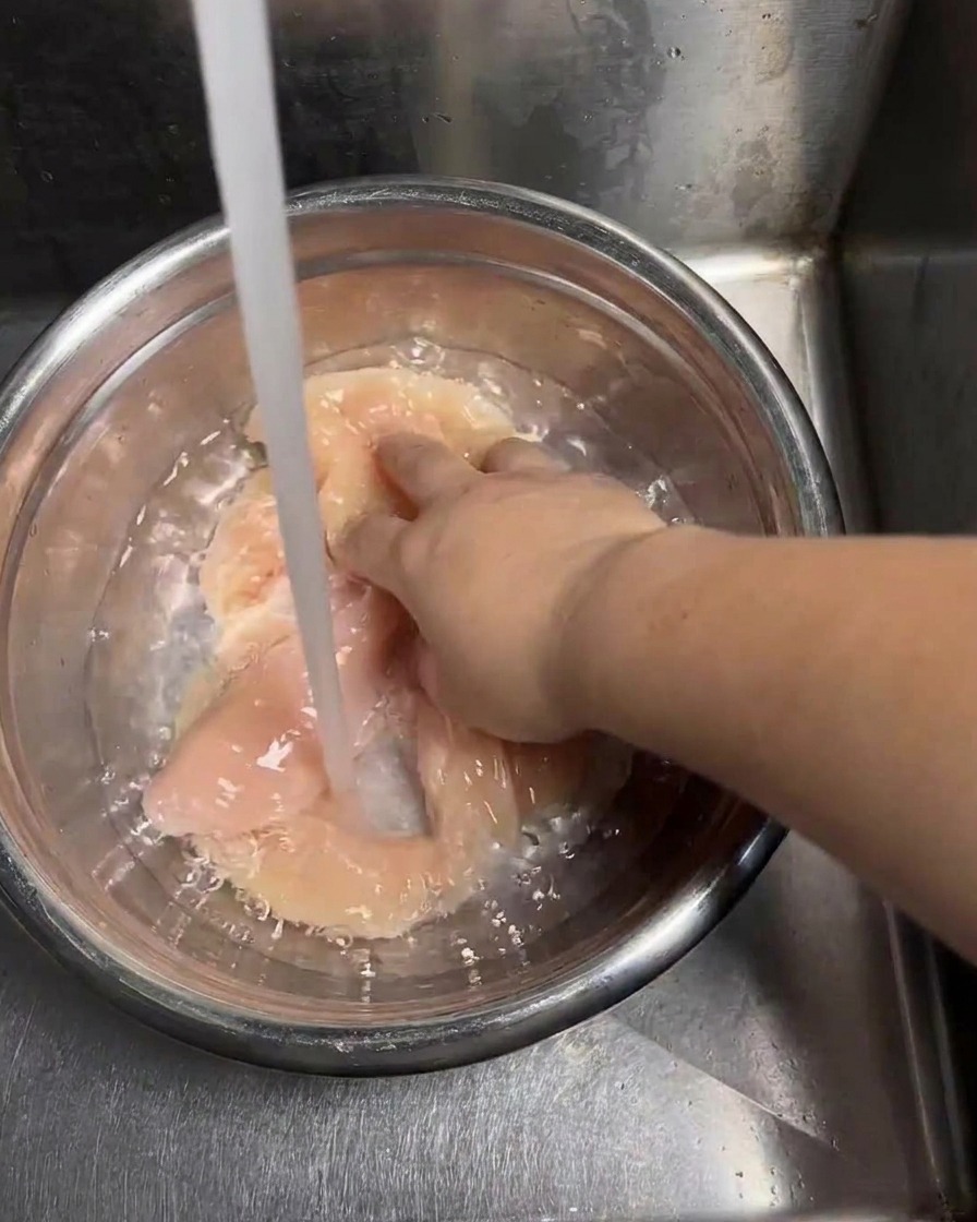 Ein Strahl kaltes Leitungswasser fließt in eine Edelstahlschüssel mit rohen Hähnchenschnitzeln, während eine Hand die Stücke zum Waschen wendet.