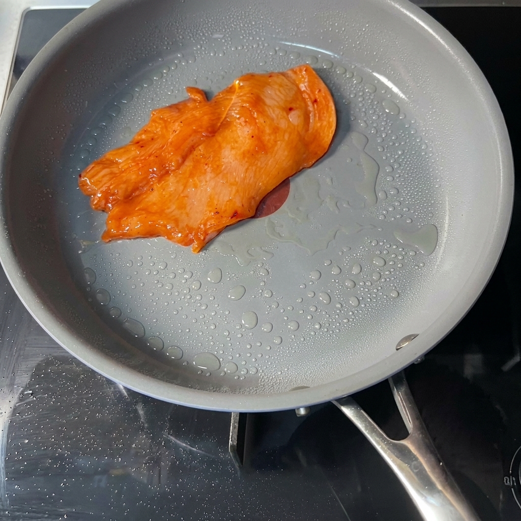 Ein einzelnes, mariniertes, orangefarbenes Hähnchenschnitzel brät in einer grauen, beschichteten Pfanne mit sichtbaren Öltropfen auf der Oberfläche.