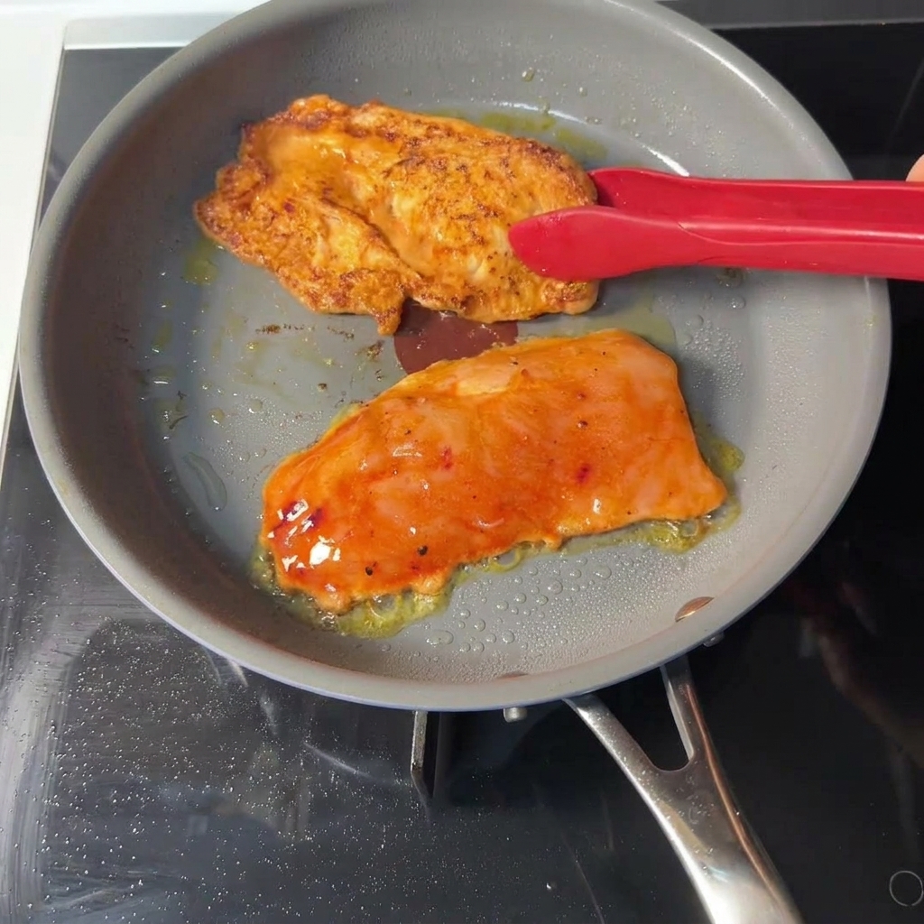 Eine rote Küchenzange wendet ein orangefarbenes, mariniertes Hähnchenschnitzel in einer grauen Pfanne und gibt den Blick auf eine goldbraun gebratene Kruste frei.