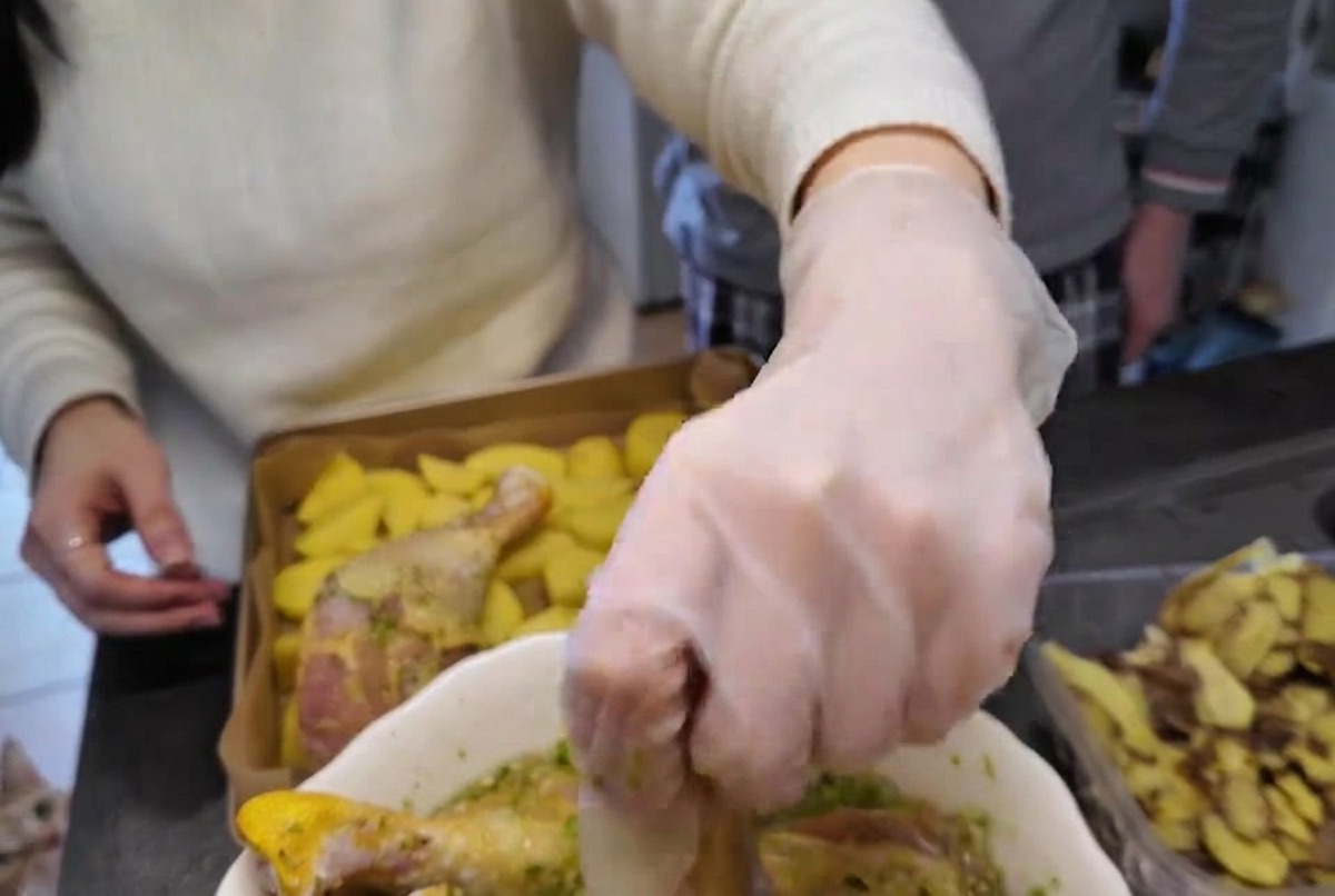 Eine behandschuhte Hand hebt einen rohen, mit grüner Kräutermarinade überzogenen Hähnchenschlegel aus einer weißen Schüssel.