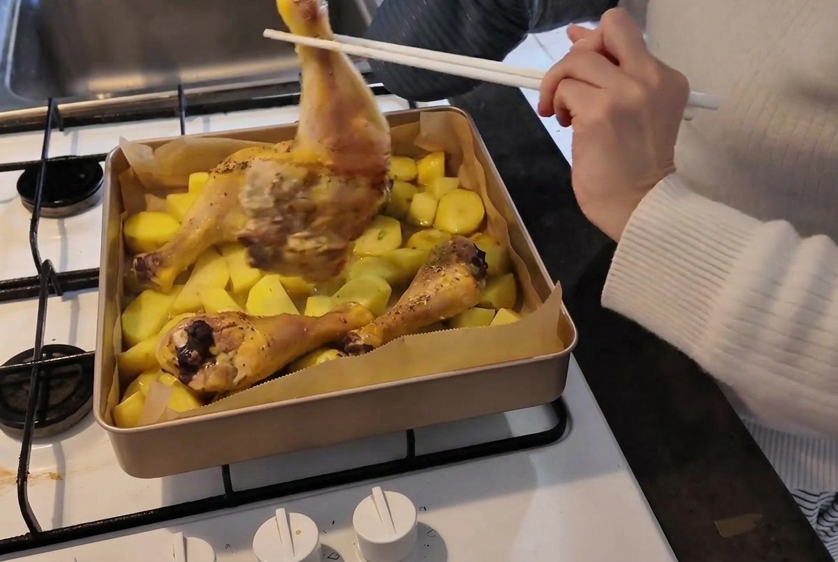 Eine Person mit Ofenhandschuh wendet einen teilweise gegarten Hähnchenschenkel mit Essstäbchen auf einem heißen Backblech voller Kartoffeln.