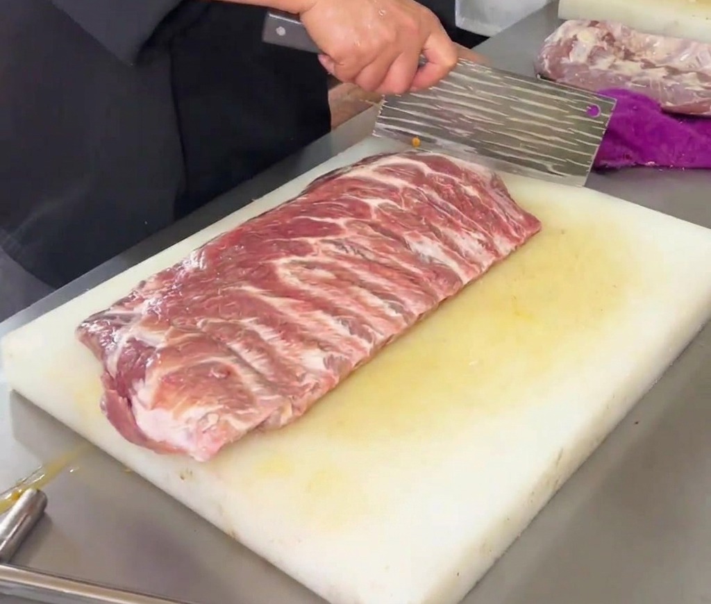 Ein Koch schneidet mit einem großen Küchenbeil ein Rautenmuster in ein rohes Stück Schweinerippchen auf einem weißen Kunststoff-Schneidebrett.