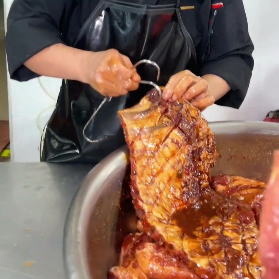 Ein Koch sticht einen großen Metallhaken in die obere Ecke eines vollständig marinierten Stücks Schweinerippchen mit dunkler Sauce über einer Edelstahlschüssel.
