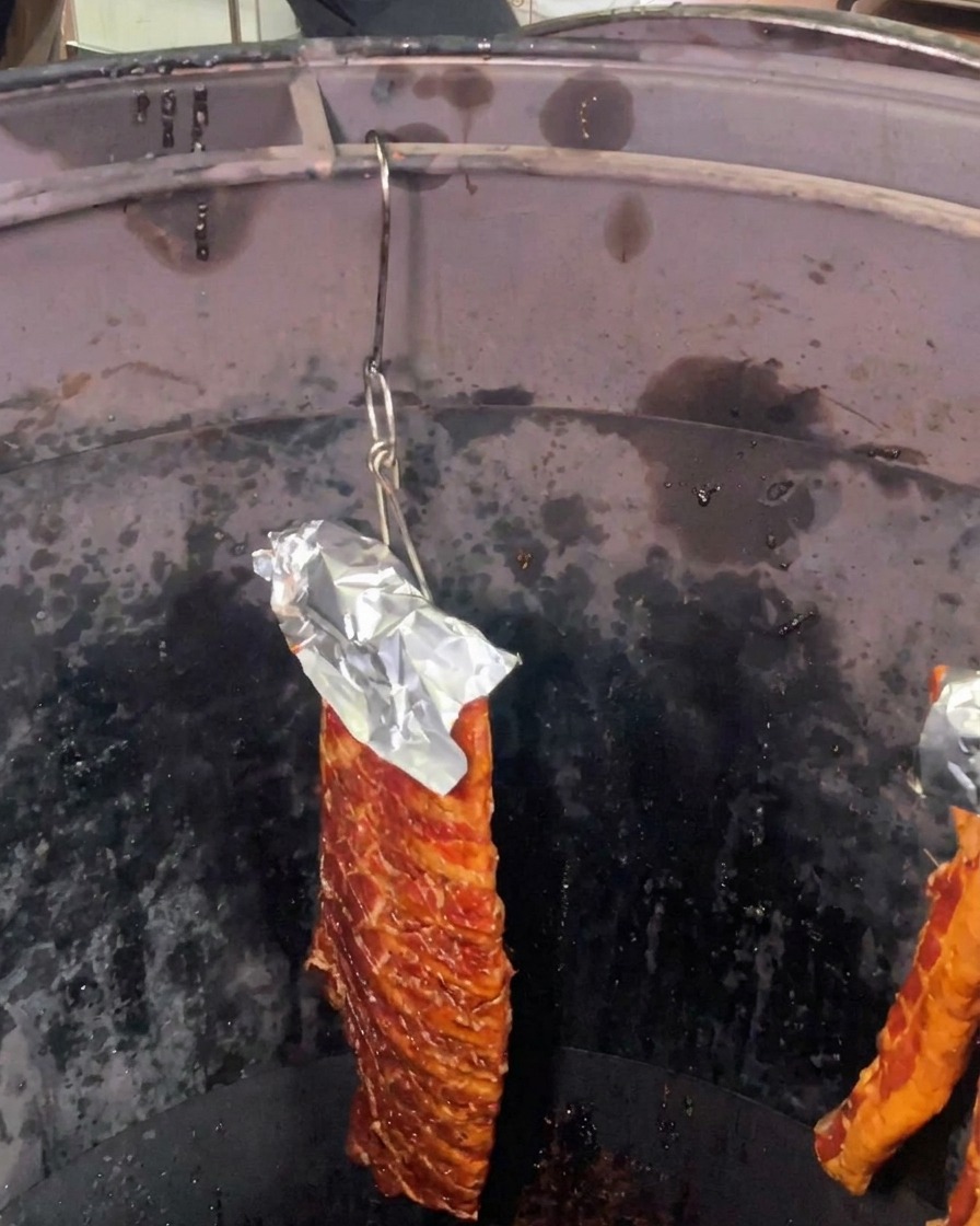 Ein Stück marinierte Schweinerippchen, deren Oberseite mit Alufolie geschützt ist, hängt in einem tiefen, dunklen, zylindrischen Röstofen.
