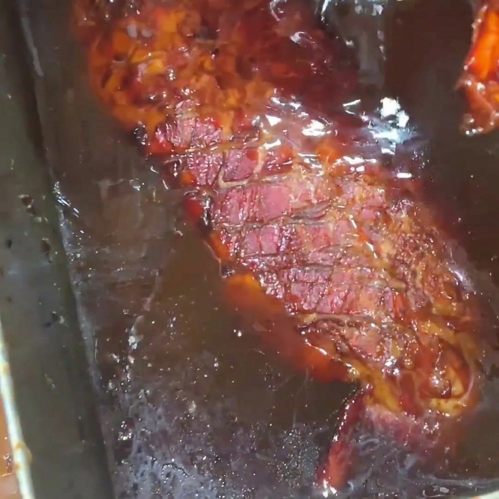 Ein goldbraun geröstetes Stück Schweinerippchen wird in einer Edelstahlschale mit einer reichhaltigen, glänzenden Honigsirup-Glasur übergossen.
