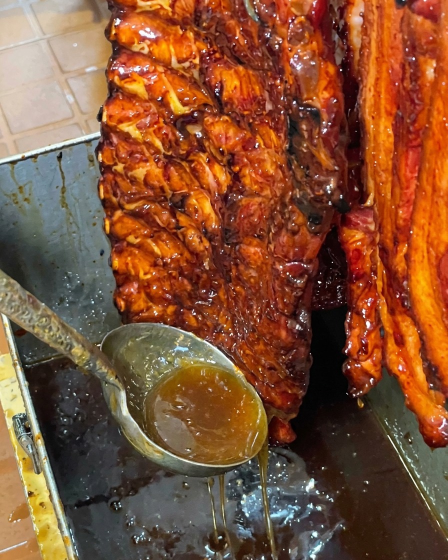 Ein glänzendes, honigglasiertes Stück Schweinerippchen hängt vertikal über einem Metallbecken, während überschüssige süße Glasur herabtropft.