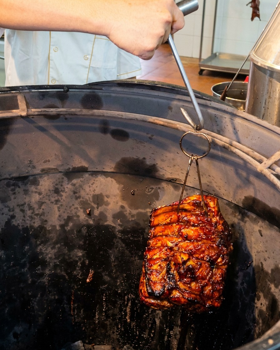Ein Stück glänzende Schweinerippchen wird mit einem Metallhaken in einen tiefen, dunklen, zylindrischen Röstofen herabgelassen.