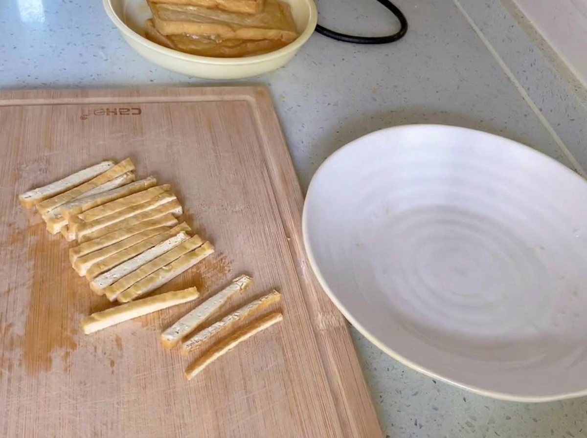 Goldbraun frittierter Tofu, in gleichmäßige dünne Streifen geschnitten, auf einem Holzschneidebrett neben einem weißen Teller.