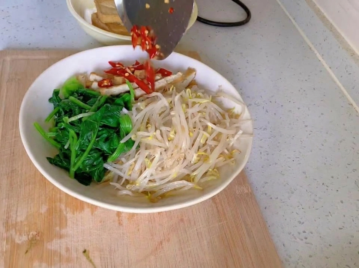Eine weiße Schüssel mit getrennten Bereichen aus grünem Spinat, hellen Sprossen und Tofustreifen, getoppt mit gehackten roten Chilis und feingehacktem Knoblauch.