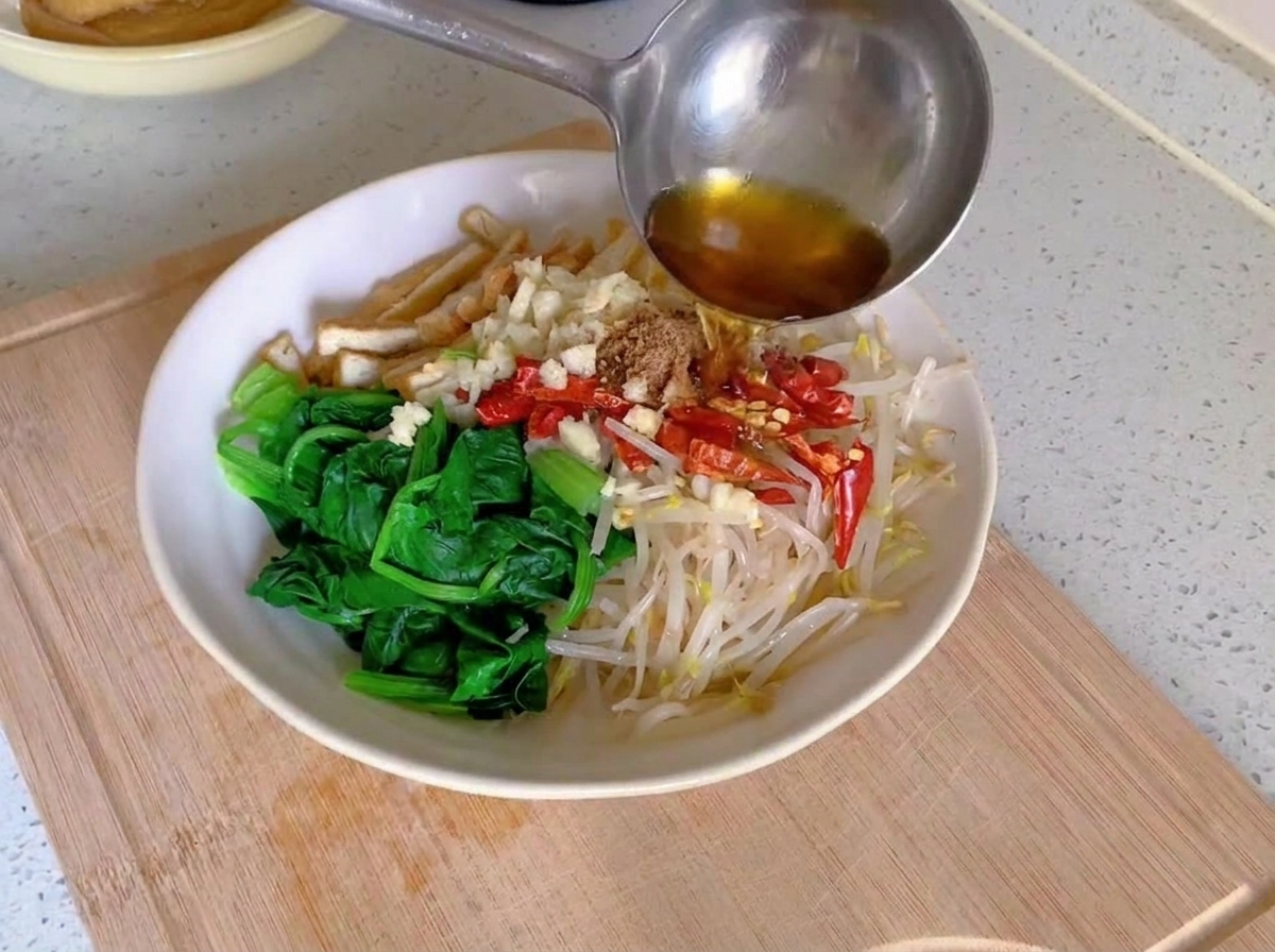 Rauchend heißes Öl wird aus einer Metallkelle über gehackten Knoblauch und rote Chilis gegossen, die auf blanchiertem Gemüse in einer weißen Schüssel liegen.