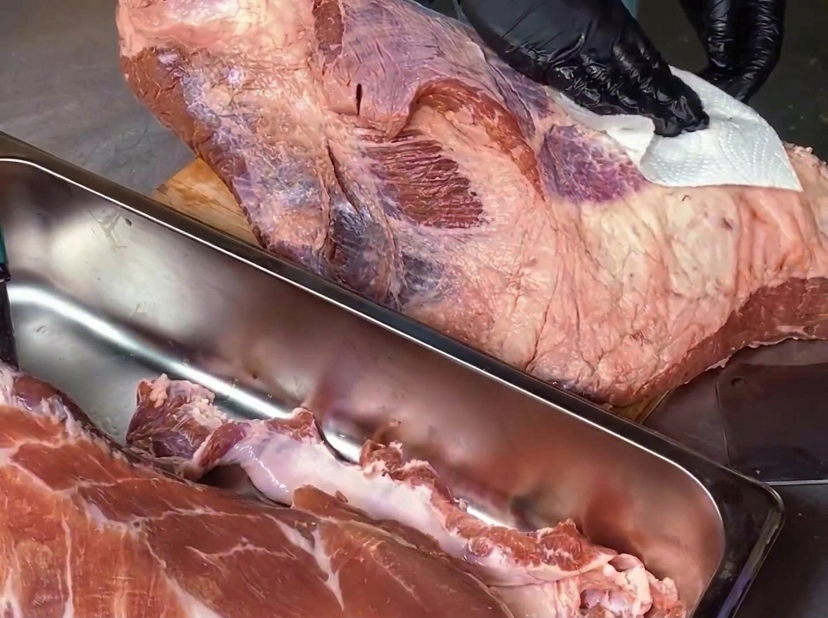 Ein Koch mit schwarzen Handschuhen tupft ein großes Stück rohe Rinderbrust auf einem Holzschneidebrett mit einem weißen Papiertuch ab, im Vordergrund steht eine Edelstahlschale mit Abschnitten.