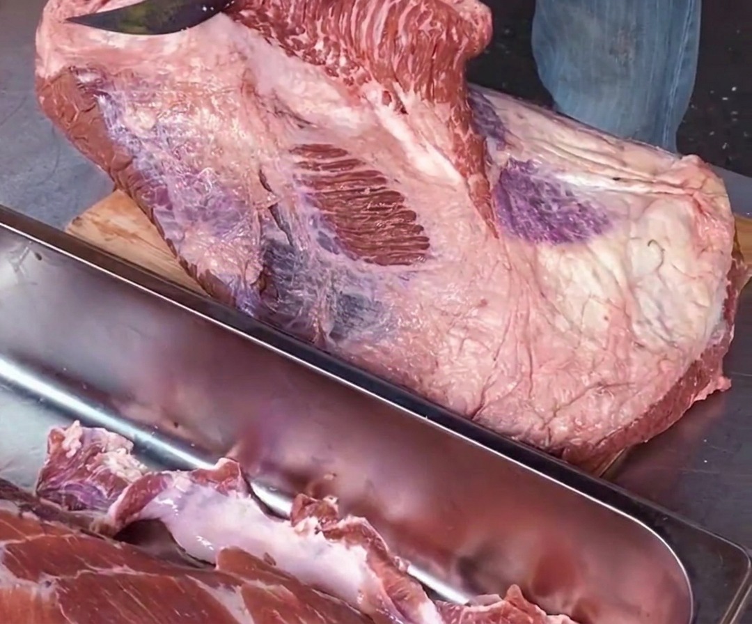 Nahaufnahme eines Kochs, der mit einem scharfen Messer parallel zum Schneidebrett das rohe Point-Fleisch entlang einer weißen Fettnaht vom Flat-Fleisch trennt.