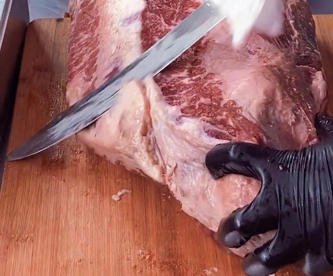 Eine Hand in einem Handschuh stabilisiert ein Stück Rinderbrust, während ein langes Messer eine dicke Schicht weißen Fettes vom roten Fleisch wegschneidet.