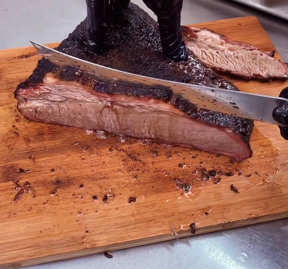 Ein Koch schneidet ein perfekt geräuchertes Beef Brisket mit dunkler Kruste gegen die Faser in dünne Scheiben auf einem Holzschneidebrett.