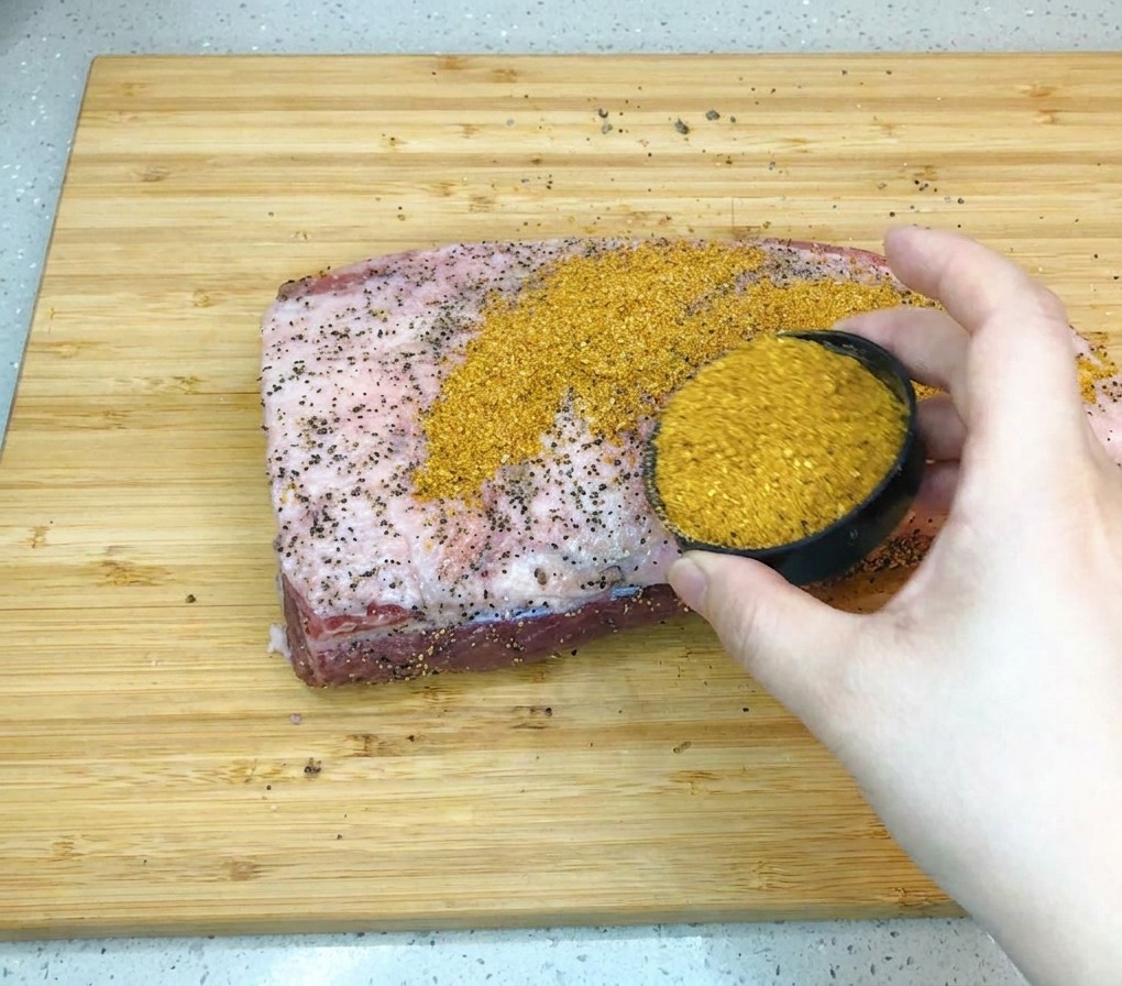 Eine Hand gießt die goldbraune Barbecue-Gewürzmischung aus einer kleinen Schale auf ein großes Stück rohe Rinderbrust, die auf einem Holzbrett liegt.