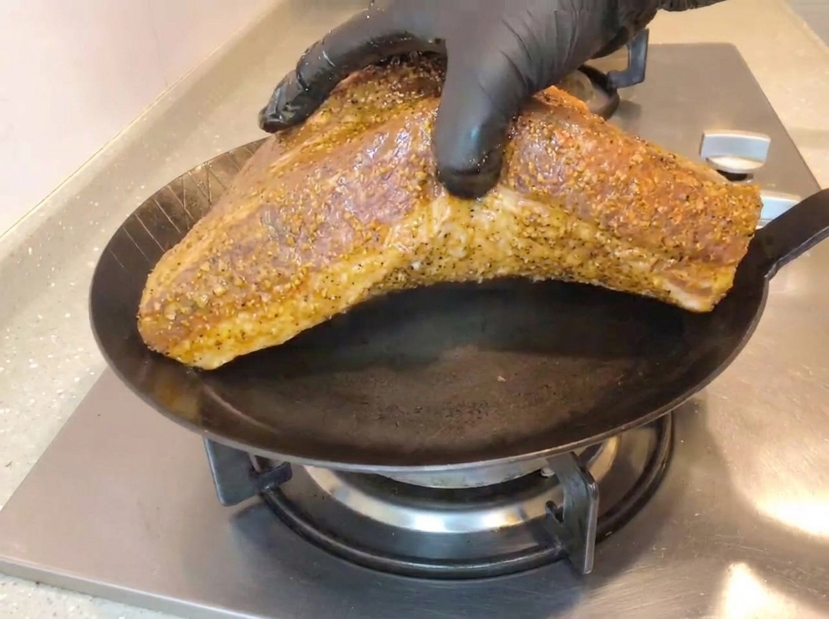 Eine Hand mit einem schwarzen Handschuh legt die große Rinderbrust mit der Fettseite nach unten in eine heiße, gusseiserne Pfanne auf dem Gasherd.