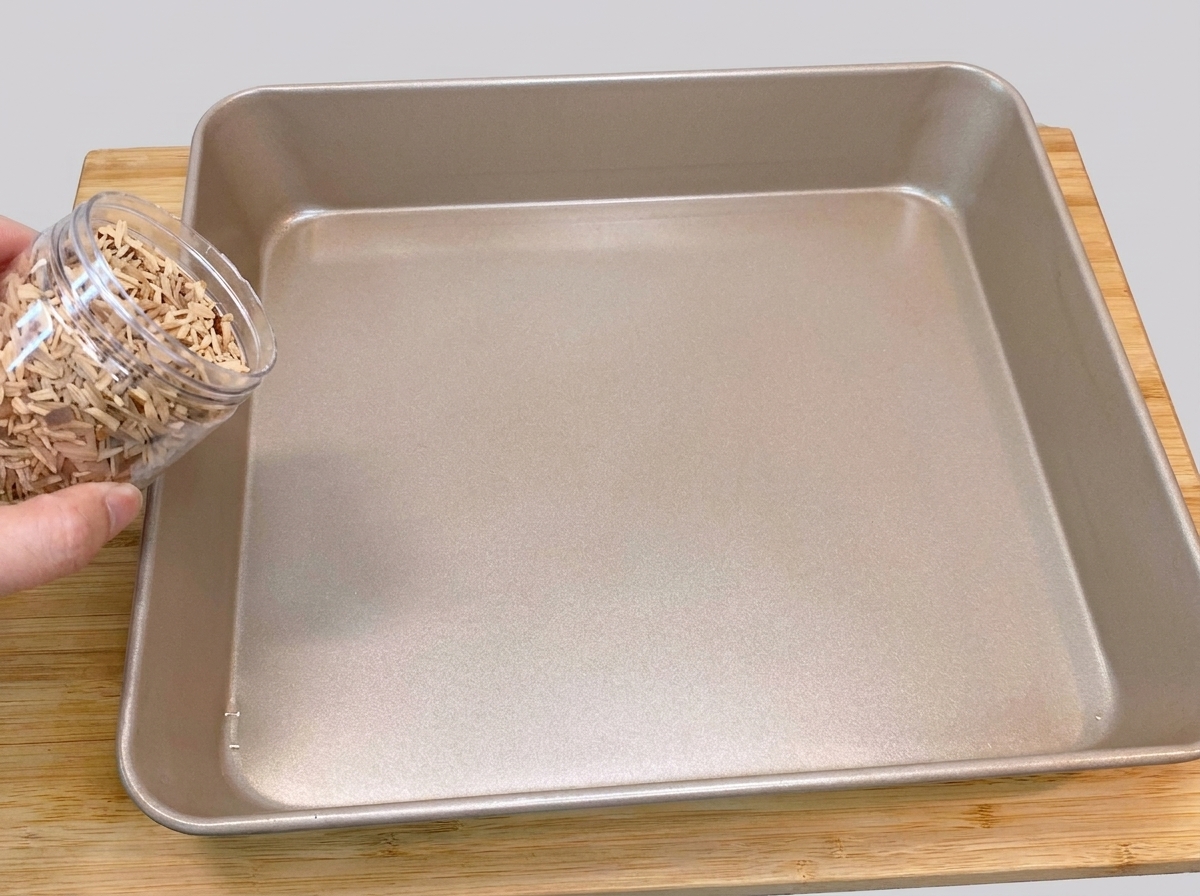 Eine Hand schüttet Pfirsichholz-Räucherchips aus einem Plastikbehälter auf den Boden einer quadratischen, goldfarbenen Backform.