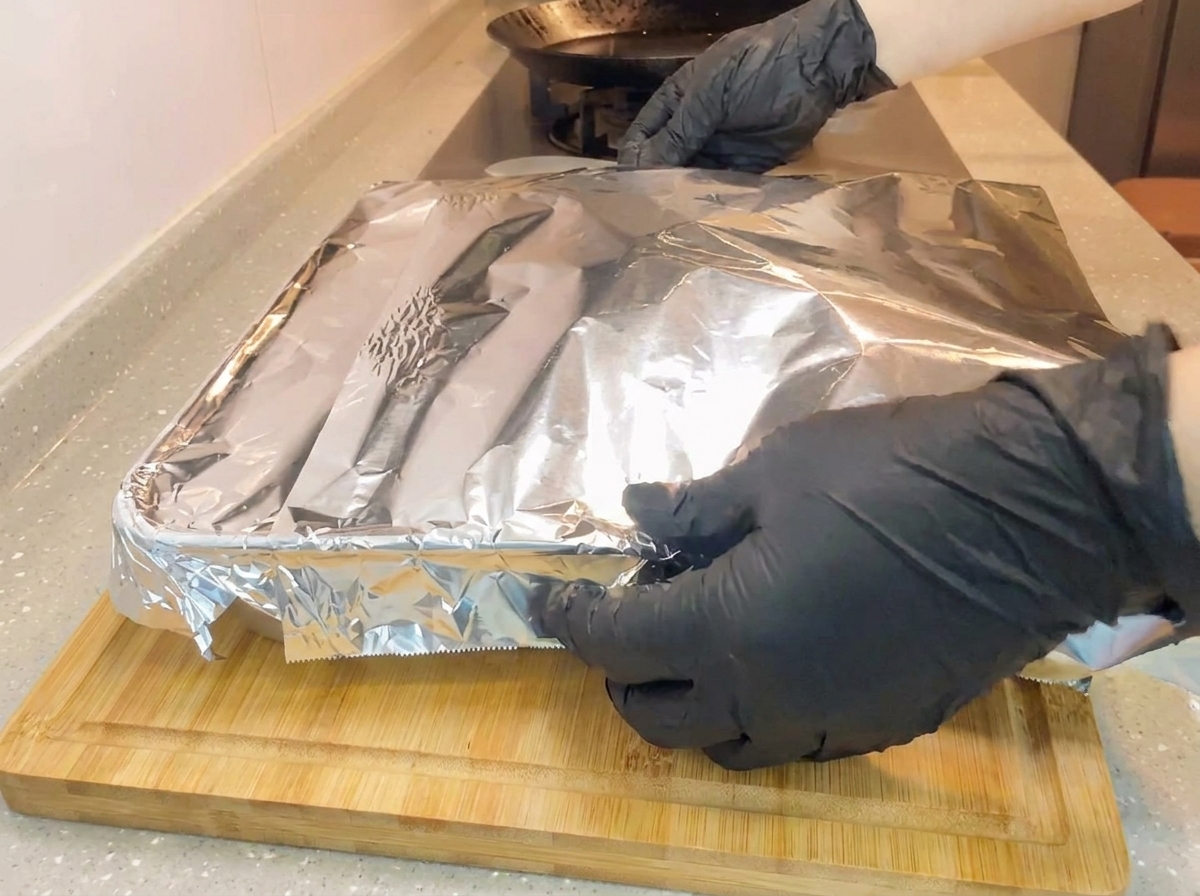 Hände mit Ofenhandschuhen wickeln eine Schicht Alufolie fest um eine quadratische Backform auf einem Holzschneidebrett.