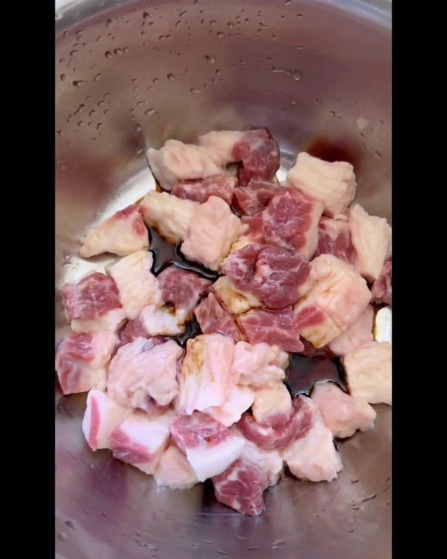 Dunkle Sojasauce und flüssige Gewürze sammeln sich um die rohen Würfel aus Rinderbrustfett in einer Edelstahlschüssel.
