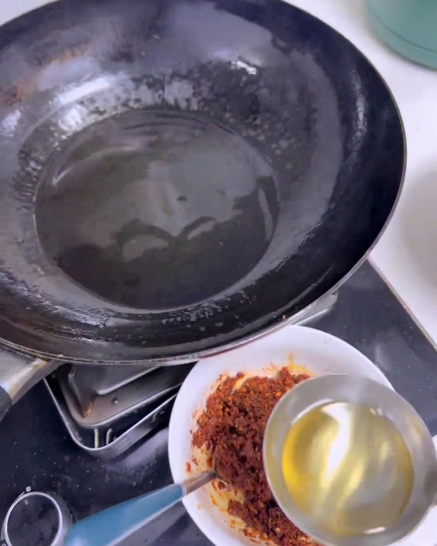 Eine Edelstahlkelle gießt klares, heißes Öl in eine weiße Schüssel mit einer dunkelroten Mischung aus Chilipulver und Gewürzen neben einem schwarzen Wok.