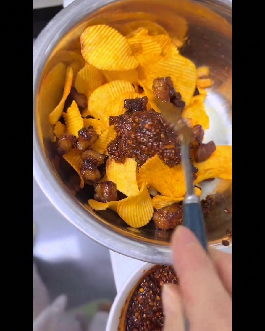 Eine Hand mischt mit einem Löffel Riffel-Kartoffelchips, knuspriges Rinderfett und leuchtend rotes Chiliöl in einer Metallschüssel.