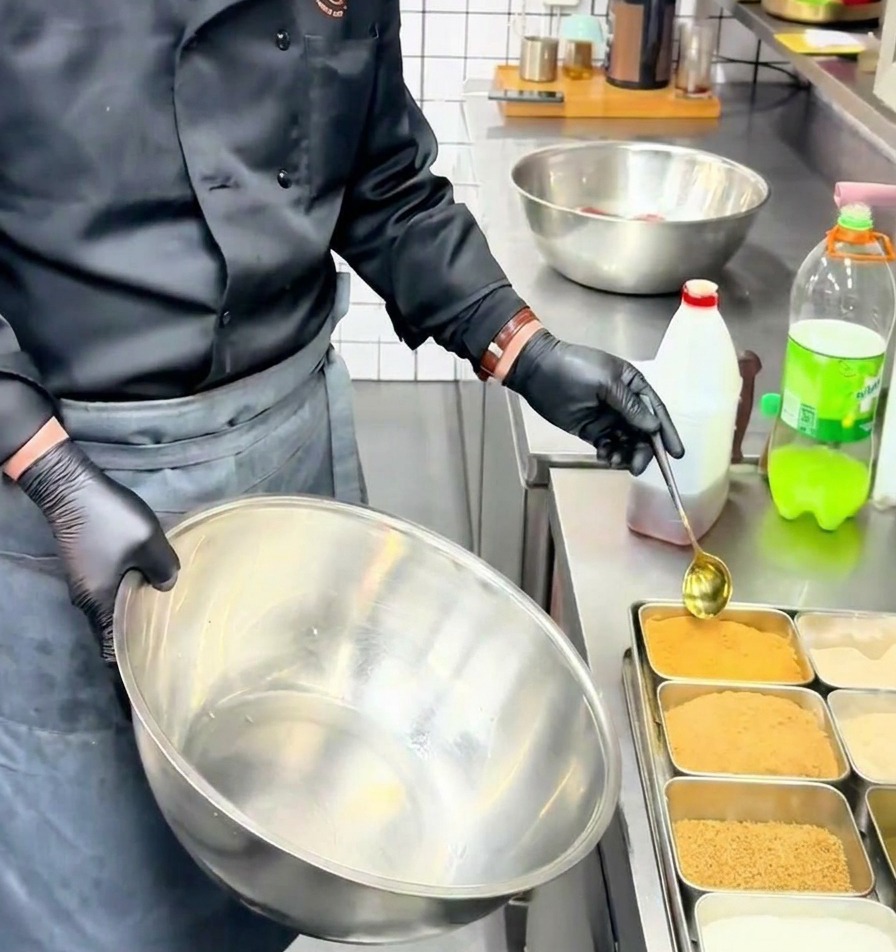 Ein Koch gibt mit einem goldenen Löffel gelbes Rinderbrühe-Pulver in eine große Rührschüssel aus Edelstahl.