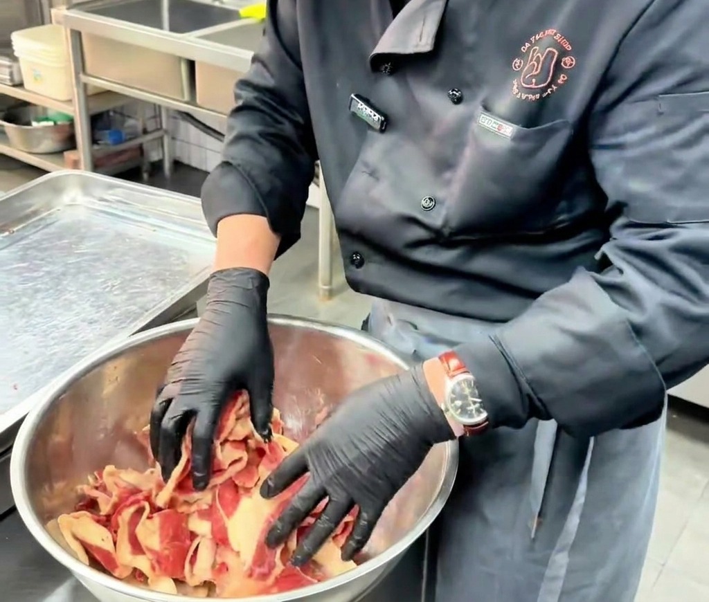 Ein Koch vermengt mit den Händen in schwarzen Handschuhen dünne, rohe Rindfleischscheiben mit der Marinade in einer großen Edelstahlschüssel.