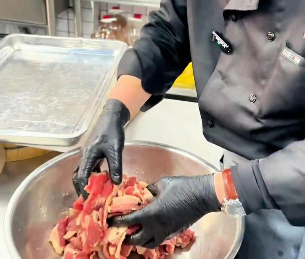 Hände in Handschuhen wenden dünne Rindfleischscheiben mit Öl in einer Metallschüssel.