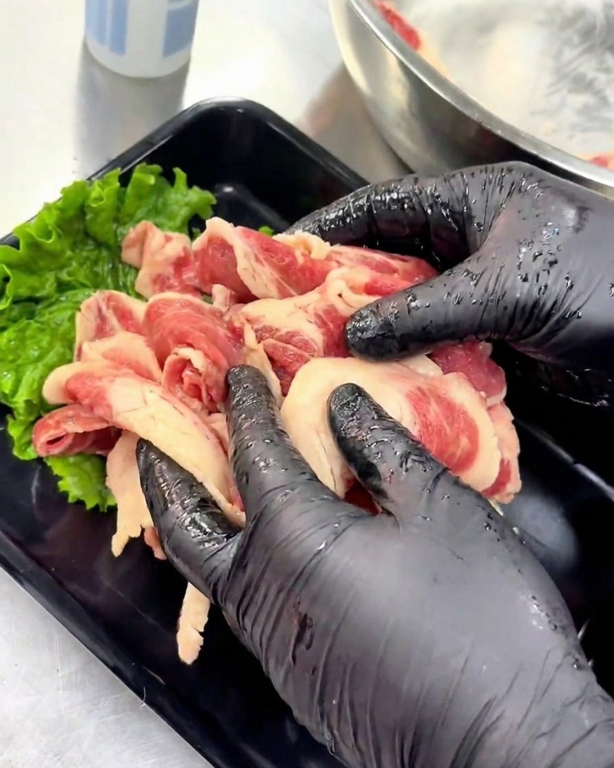 Hände in Handschuhen richten marinierte rohe Rindfleischscheiben ordentlich auf einem schwarzen Serviertablett an, das mit einem grünen Salatblatt dekoriert ist.