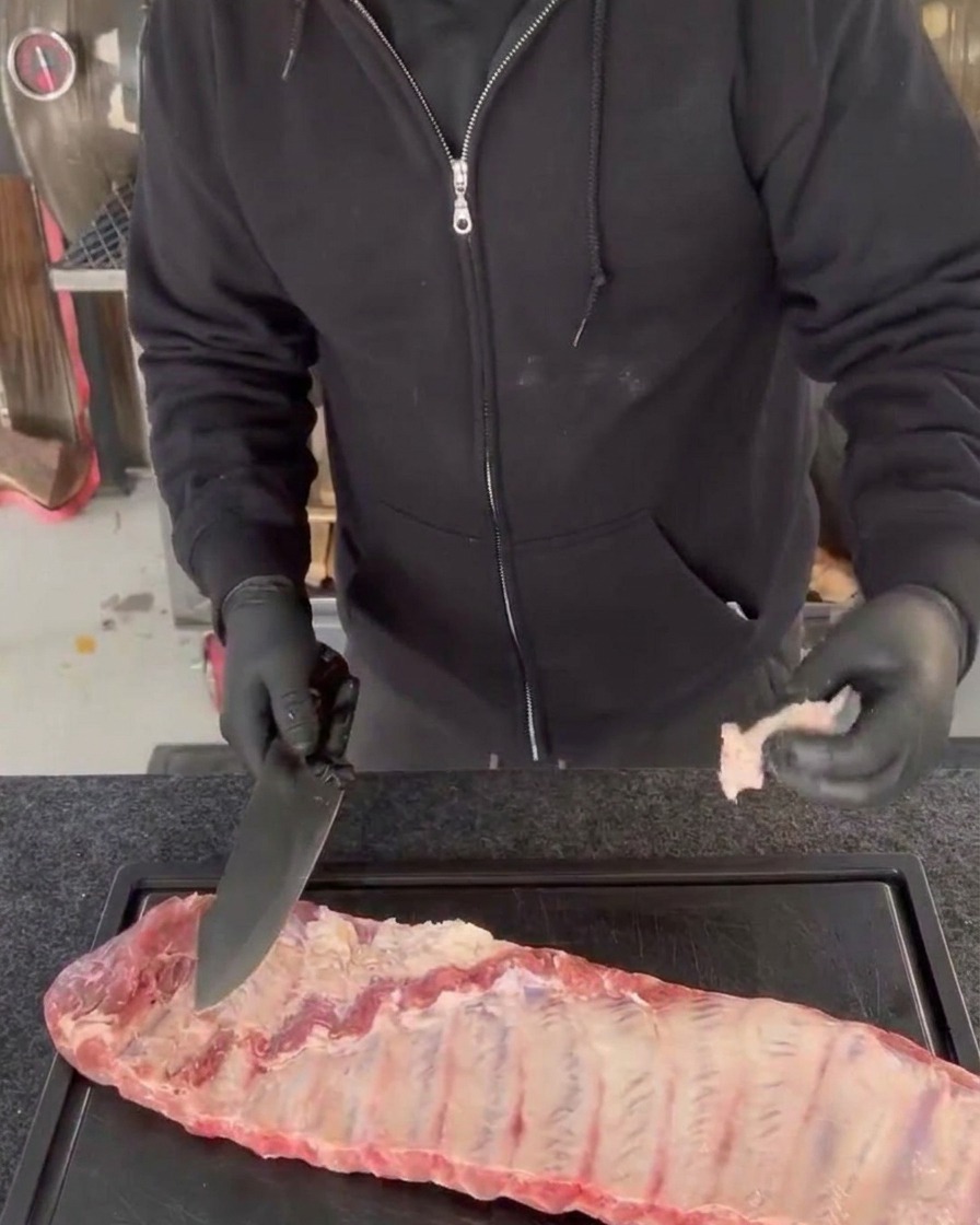 Eine Person mit schwarzen Handschuhen schneidet überschüssiges Fleisch und unebene Kanten von rohen Schweinerippchen ab.