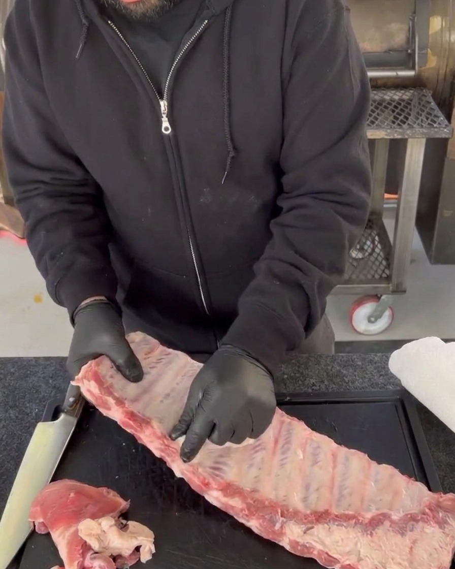 Hände mit Handschuhen ziehen die glänzende, durchsichtige Silberhaut von den Knochen roher Schweinerippchen ab.