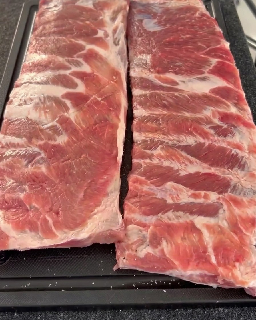 Zwei rohe Stränge Schweinerippchen nebeneinander auf einem Brett, deren Fleischseite dick mit grobem Pfeffer und Salz bedeckt ist.