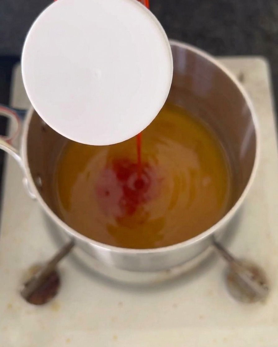 Ein Strahl roter Ketchup wird in eine köchelnde, bernsteinfarbene Flüssigkeit in einem Edelstahltopf gegossen.